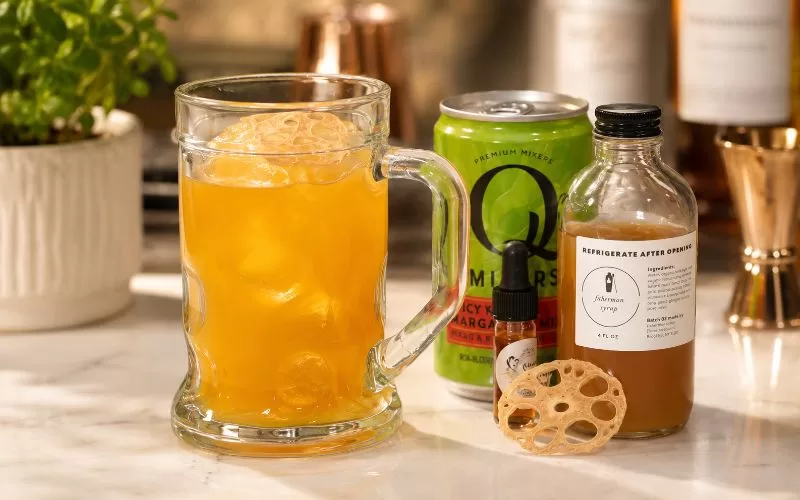 Orange craft cocktail in a glass mug with mixer can, syrup bottle, bitters, and dried lotus root garnish on a home bar counter
