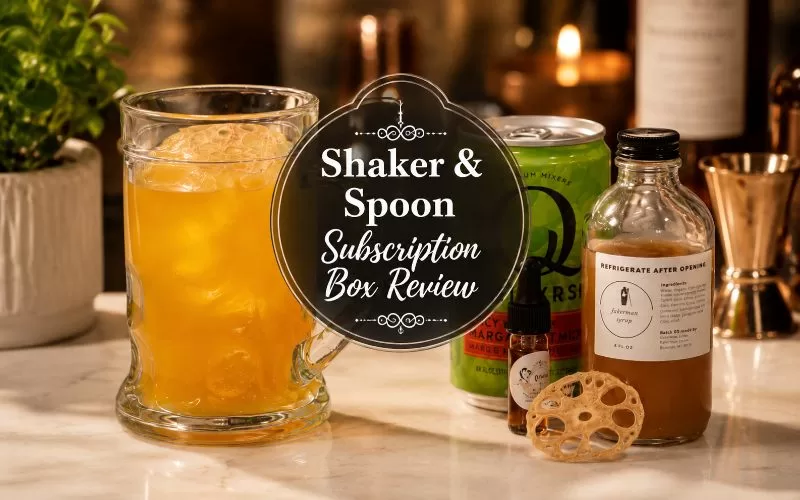 Orange craft cocktail with Shaker & Spoon mixer, syrup bottle, bitters, and dried garnish on a warm home bar counter