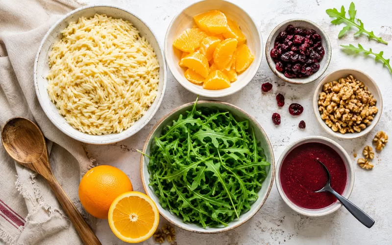 Ingredients for cranberry orange orzo salad arranged in bowls with cooked orzo, orange segments, cranberries, walnuts, arugula, and cranberry dressing