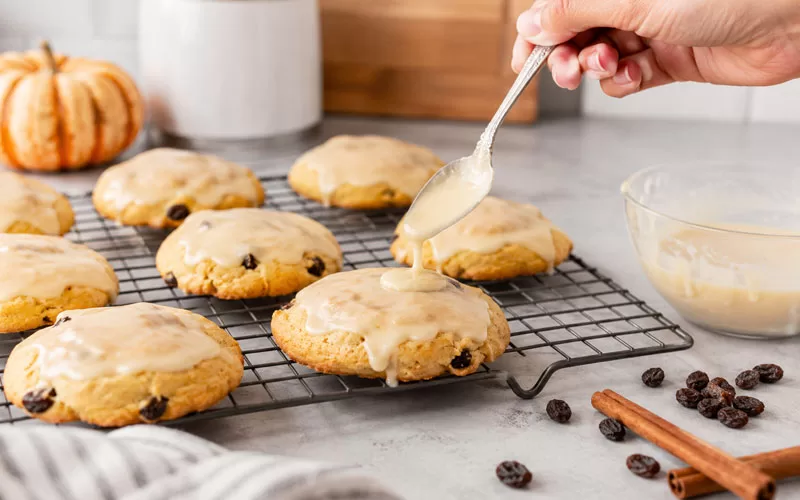 Hand spooning glaze over soft pumpkin raisin cookies on a cooling rack in a fall kitchen setting.