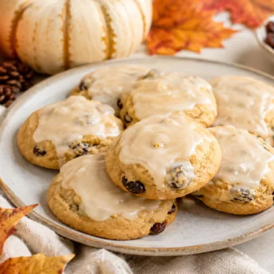 Glazed pumpkin raisin cookies on a plate with fall pumpkins, cinnamon sticks, raisins, and autumn leaves.