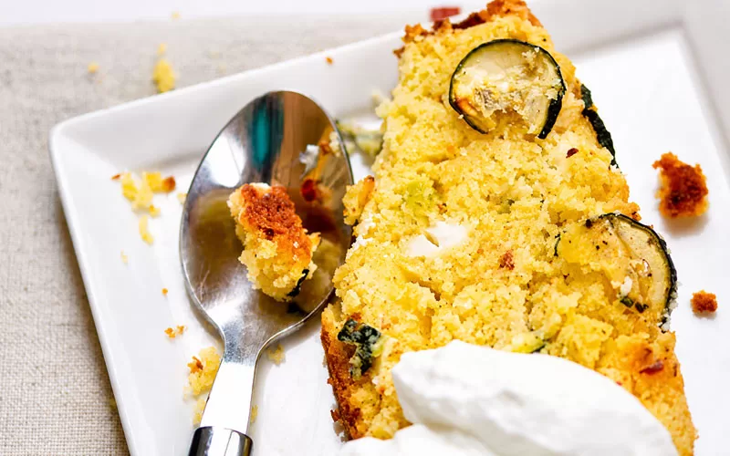 Close-up slice of zucchini feta cornbread on a white plate with a spoon and creamy topping.