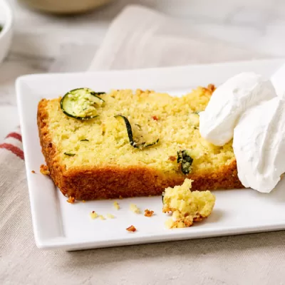 Slice of zucchini feta cornbread on a white plate with creamy topping and fresh zucchini in the background.
