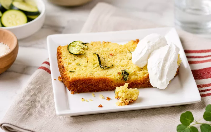 Slice of zucchini feta cornbread on a white plate with creamy topping and fresh zucchini in the background.