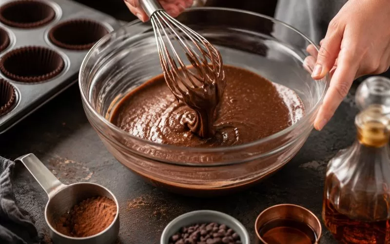 Hands whisking rich chocolate bourbon cupcake batter in a glass mixing bowl beside baking ingredients and cupcake liners.