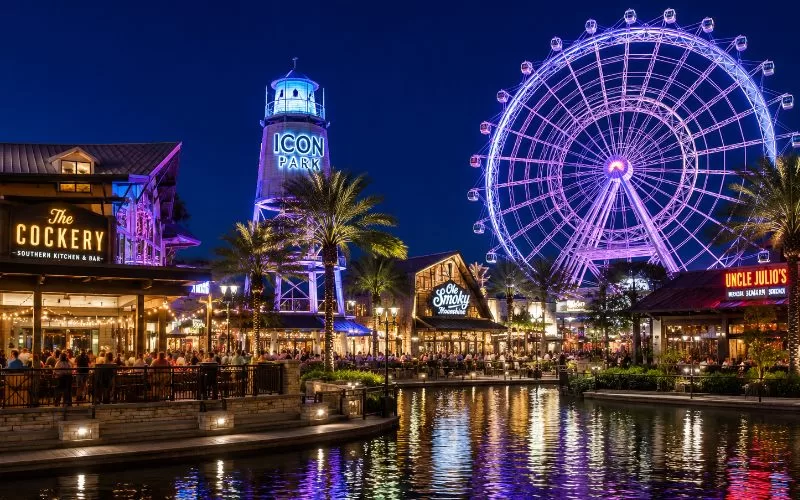 Nighttime Orlando entertainment district with glowing lights, waterfront restaurants, palm trees, and a large illuminated observation wheel at ICON Park.