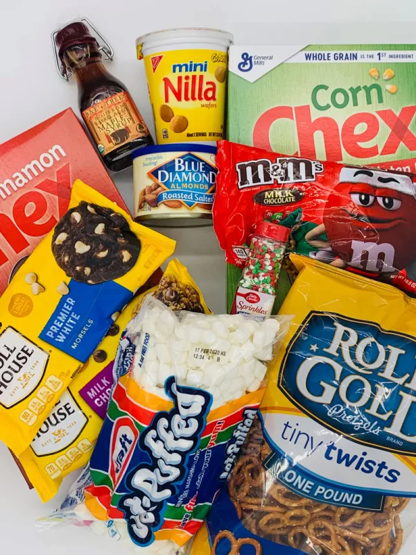 Flat lay of ingredients for Festive Sweet Maple Christmas Chex Mix including Corn Chex cereal, pretzels, marshmallows, vanilla wafers, almonds, maple syrup, chocolate chips, M&Mโs, and holiday sprinkles.