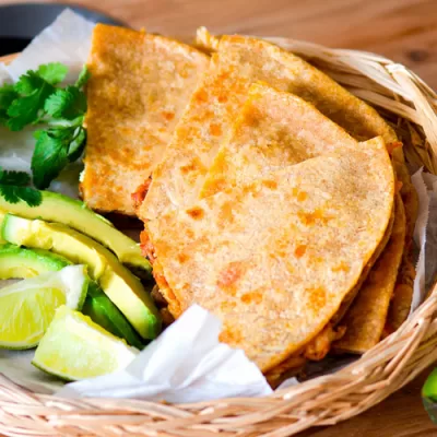 Basket of crispy rotisserie chicken quesadillas served with fresh avocado slices, lime wedges, and cilantro on a wooden table.