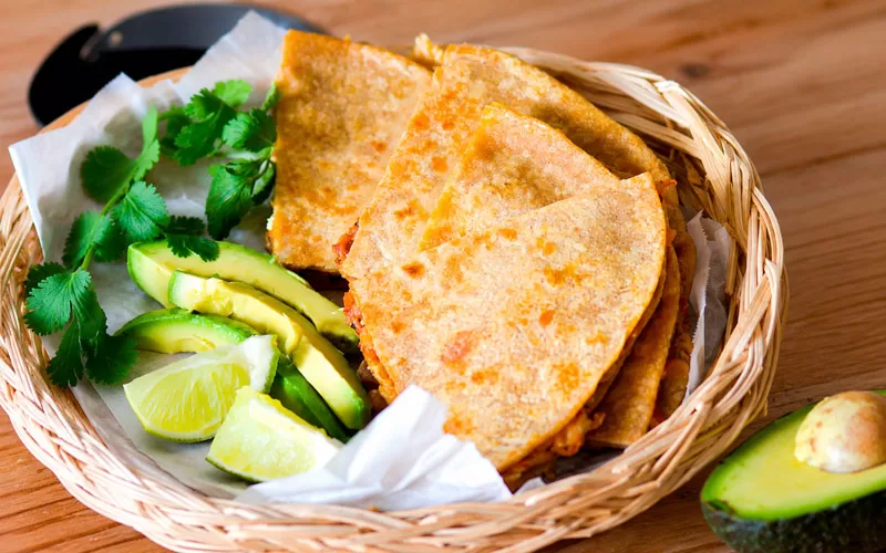 Basket of crispy rotisserie chicken quesadillas served with fresh avocado slices, lime wedges, and cilantro on a wooden table.