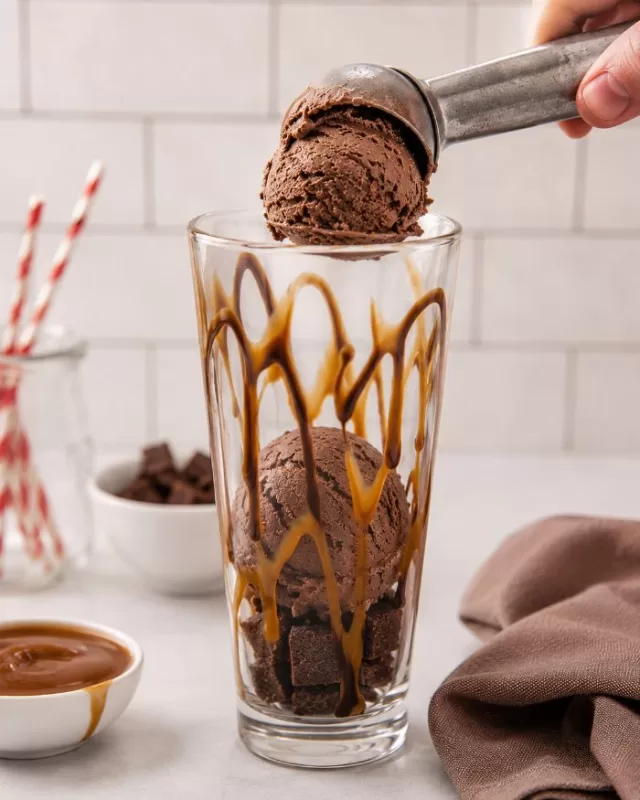 Chocolate ice cream being scooped into a tall float glass drizzled with chocolate and caramel sauce over brownie pieces.