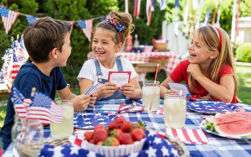 Three children laughing together while reading Fourth of July joke cards at a patriotic picnic table with lemonade and small American flags.