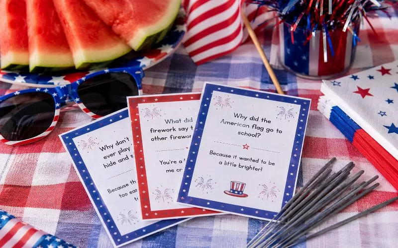 Fourth of July joke cards arranged beside sparklers, patriotic sunglasses, watermelon, American flags, and festive table decorations.