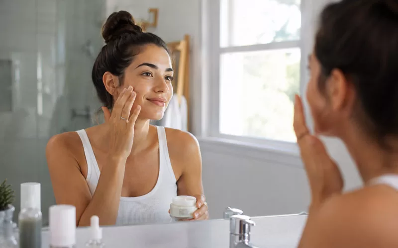 Woman applying moisturizer at a bright bathroom vanity as part of her daily skincare routine
