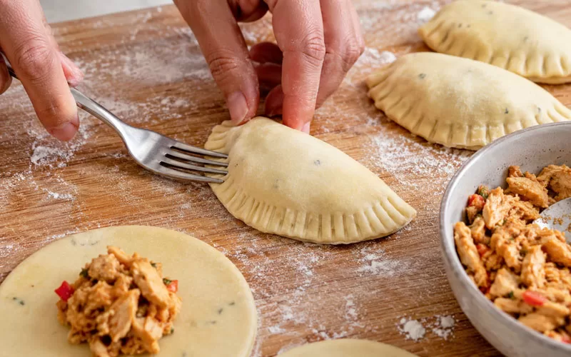 Hands sealing a homemade chicken empanada with a fork on a floured wooden board with chicken filling nearby.