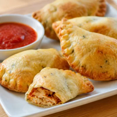 Golden chicken empanadas served on a white plate with red dipping sauce and one empanada opened to show the chicken filling.