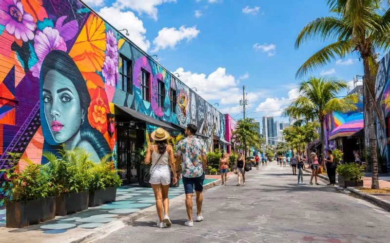 Colorful Wynwood-style mural street in Miami with palm trees, outdoor art, pedestrians, and bright tropical buildings.