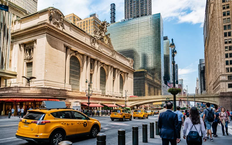 Grand Central Terminal exterior with yellow taxis, pedestrians, and classic Midtown Manhattan architecture on a bright New York City day.