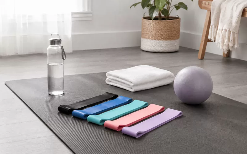 Resistance loop bands arranged on a yoga mat with a water bottle, folded towel, Pilates ball, and grey flooring in a bright home workout space.