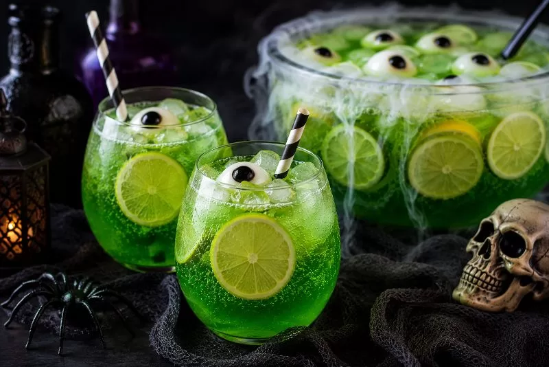 Two spooky green Halloween punch drinks with striped straws and candy eyeballs served beside a bubbling witches brew punch bowl with skull and spider decorations.