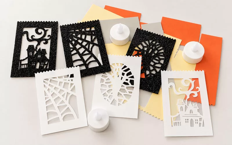 Halloween luminary craft supplies with black glitter cutouts, white cardstock panels, orange and yellow paper, and battery-operated tea lights on a neutral background.