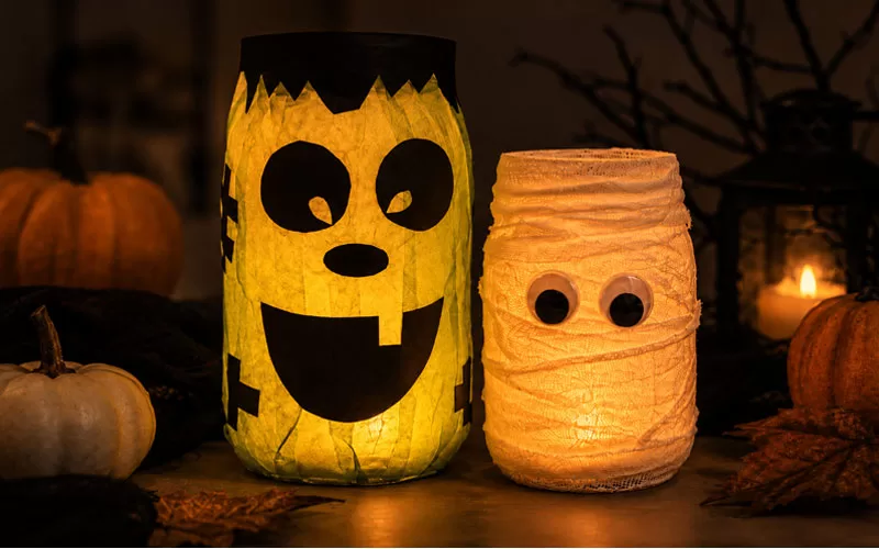 Glowing Frankenstein and mummy mason jar lanterns displayed with pumpkins and autumn leaves in a dark Halloween setting