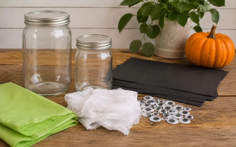 Mason jars, green tissue paper, black construction paper, gauze, and googly eyes arranged on a rustic table for a Halloween craft