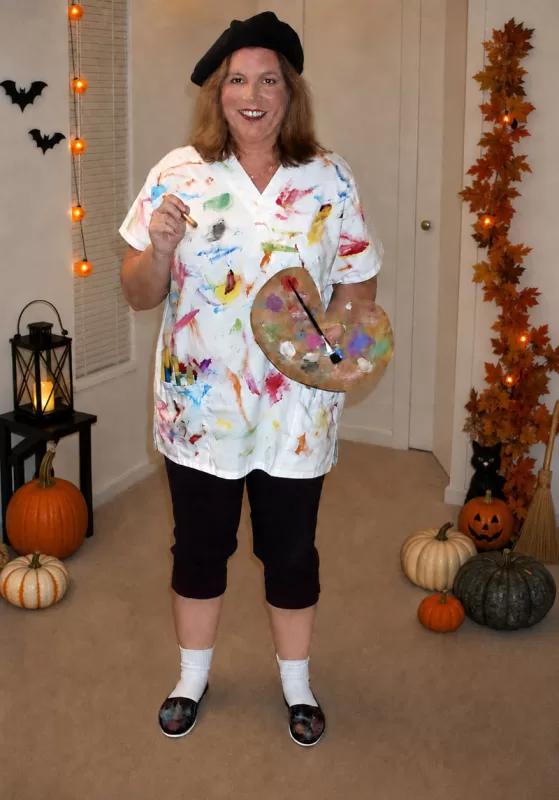 Woman dressed in a homemade painter Halloween costume holding a paint palette and brush with pumpkins, bats, and fall decorations in the background.