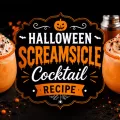 Halloween Screamsicle cocktail topped with whipped cream and black and orange sprinkles surrounded by pumpkins, spooky decor, and glowing candlelight.