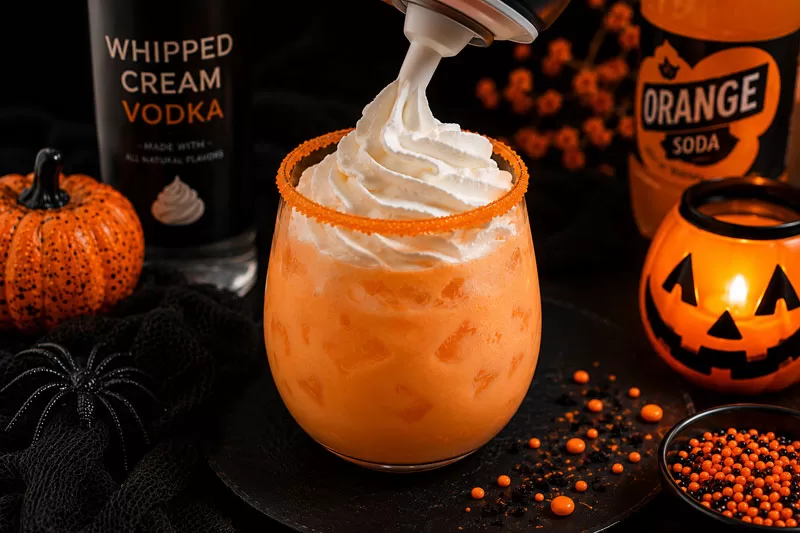 Whipped cream being added to a creamy orange Halloween Screamsicle cocktail surrounded by pumpkins, spooky decor, and glowing jack-o’-lantern candlelight.