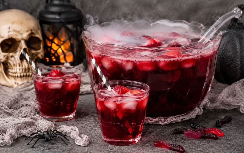 Halloween Blood Punch being poured into a glass with ice, strawberries, fog effects, gummy worms, and spooky Halloween decorations.