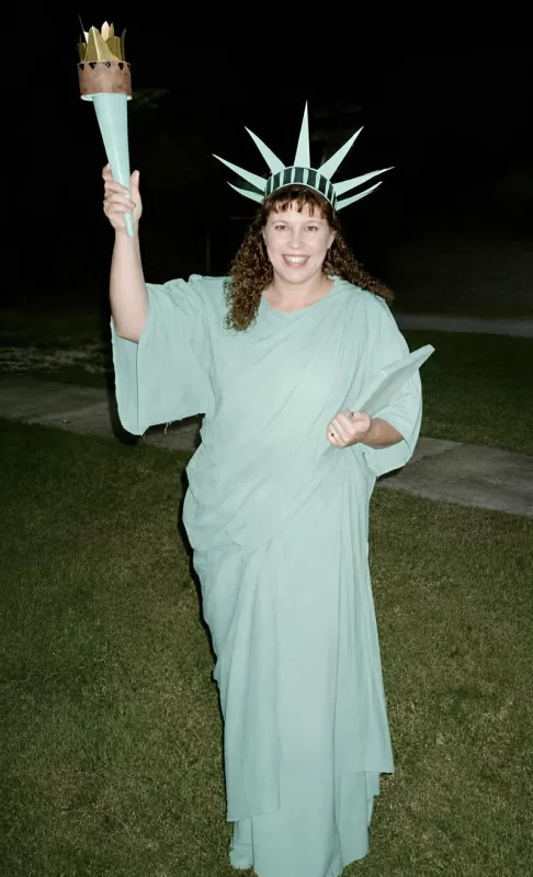 Woman dressed in a homemade Statue of Liberty costume holding a torch and tablet outside at night.