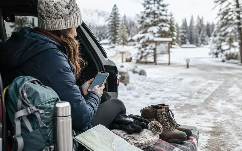 Person checking a weather map on a phone at a snowy trailhead with winter boots, gloves, backpack, thermos, and outdoor gear nearby.