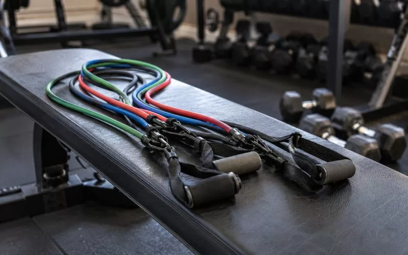Resistance bands with handles laid out on a black workout bench in a gym, with dumbbells blurred in the background.