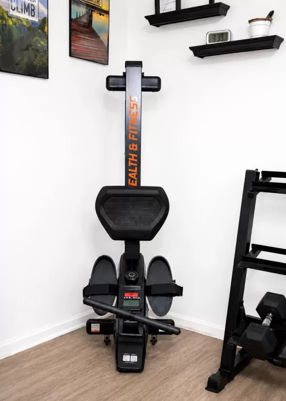 Yosuda RM2 rowing machine review image showing the compact rowing machine stored upright in a home gym corner with workout equipment, motivational wall art, and updated wood-look flooring.