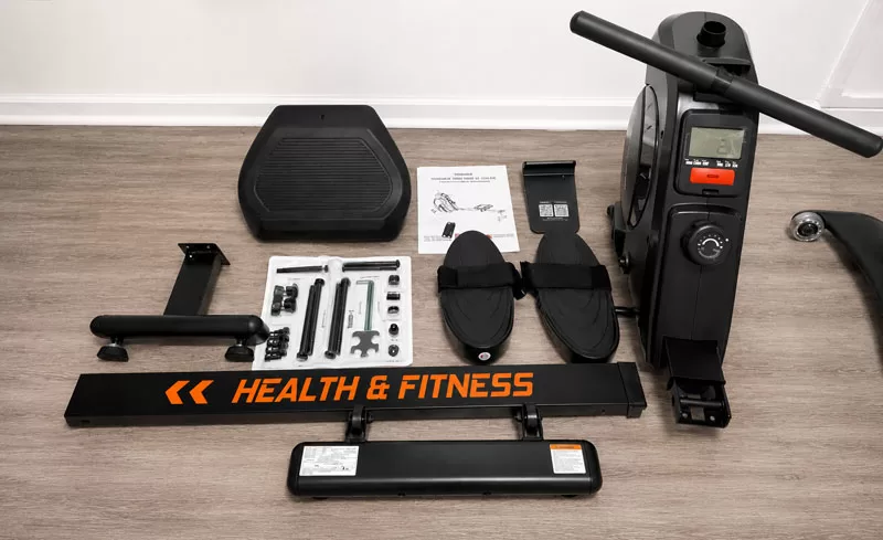 Yosuda RM2 Rowing Machine Review image showing the unassembled rowing machine parts laid out on gray wood-look flooring before setup in a home gym.