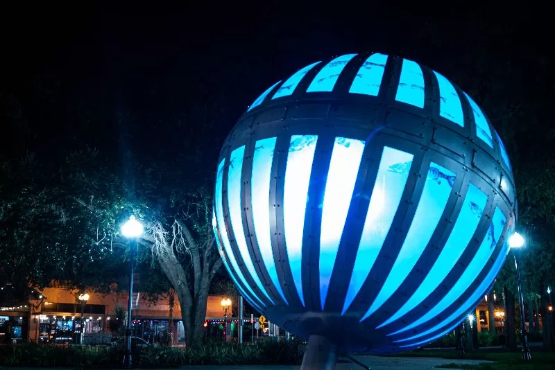 Blue illuminated Global Convergence sculpture in downtown Orlando at night surrounded by trees and glowing city lights.