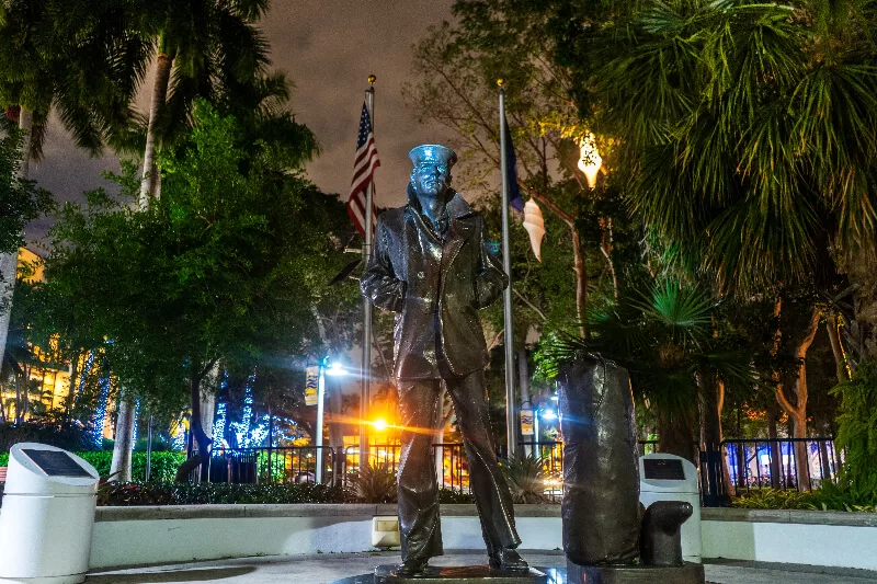 Statue along the Riverwalk in Fort Lauderdale at night surrounded by palm trees, glowing city lights, and an eerie atmosphere.