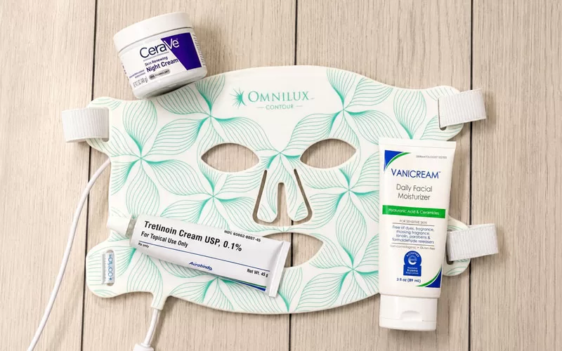 Flat lay of an over 50 skincare routine featuring an Omnilux red light therapy mask, Tretinoin cream, Vanicream moisturizer, and CeraVe night cream on a light wood background.