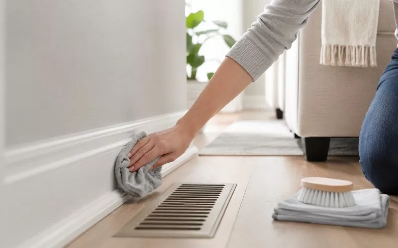 Hand wiping a white baseboard near a floor vent in a bright, neutral home.