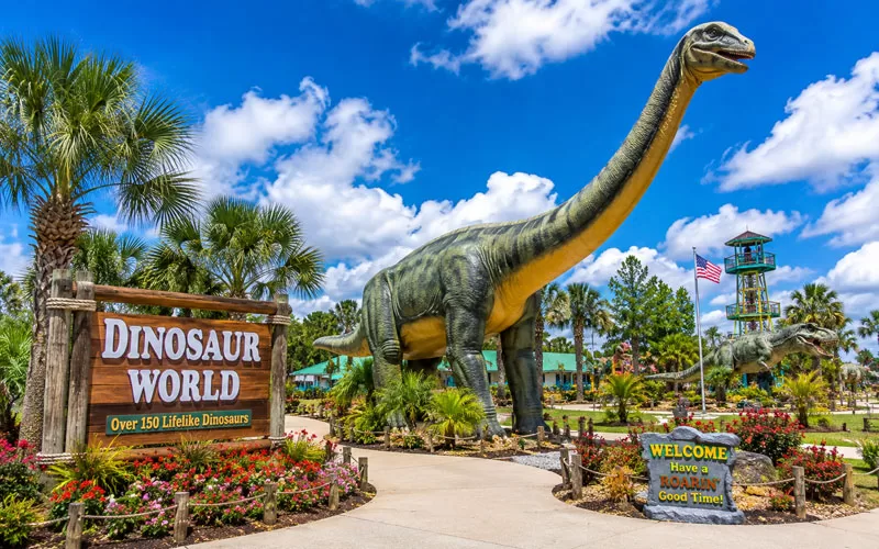 Large dinosaur statue at Dinosaur World in Florida surrounded by colorful landscaping, palm trees, and bright blue skies.