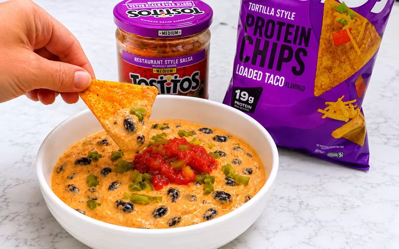 Hand dipping a Quest loaded taco protein chip into a creamy cottage cheese queso dip topped with black beans, salsa, and diced jalapeños.