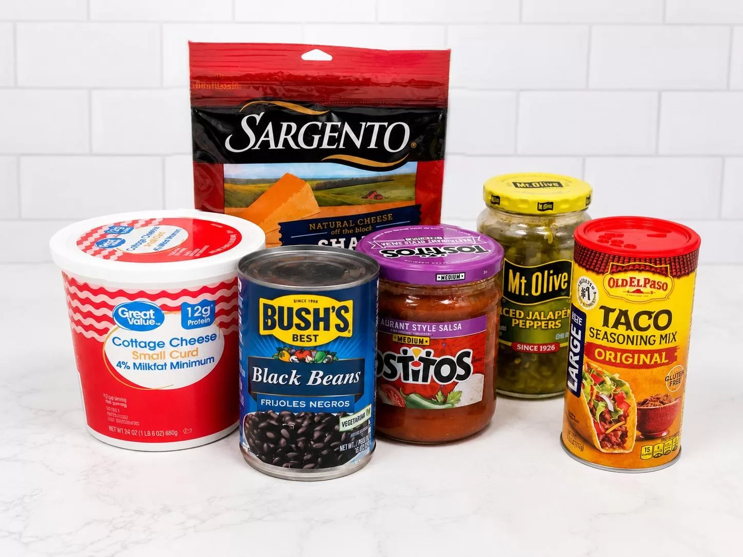 Ingredients for high protein cottage cheese queso including cottage cheese, shredded cheddar cheese, black beans, salsa, jalapeños, and taco seasoning arranged on a white countertop.