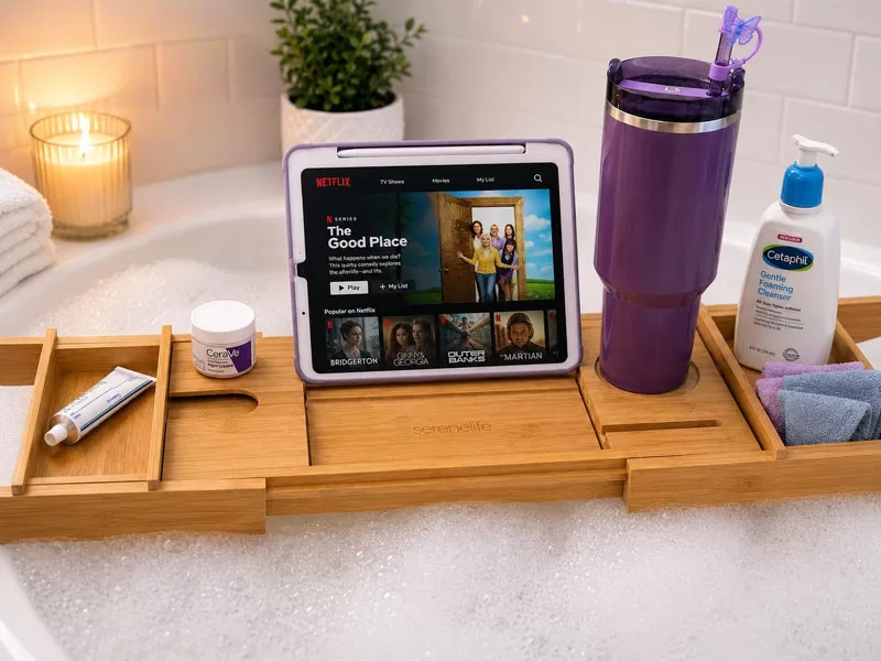 Cozy nighttime skincare setup with a wooden bath tray, skincare products, soft towels, bubbles, and a tablet beside a relaxing bubble bath.