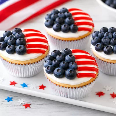 Patriotic American Flag Cupcakes decorated with fresh blueberries and red icing stripes on a white serving platter with an American flag in the background.