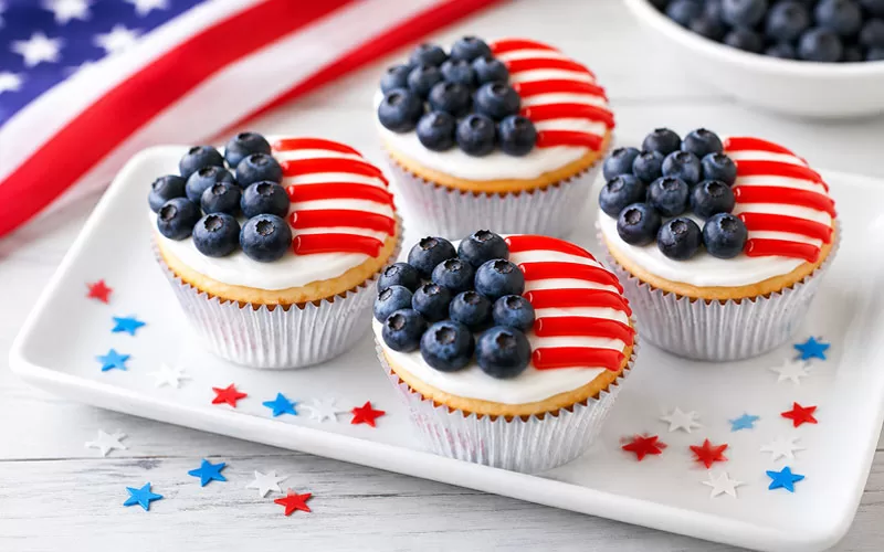 Patriotic American Flag Cupcakes decorated with fresh blueberries and red icing stripes on a white serving platter with an American flag in the background.