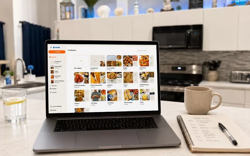Laptop displaying the ReciMe recipe dashboard on a kitchen counter with a meal planning notebook, pen, water, and coffee nearby.