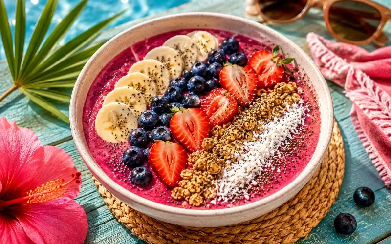 Colorful smoothie bowl topped with strawberries, blueberries, bananas, granola, coconut, and chia seeds beside a swimming pool.