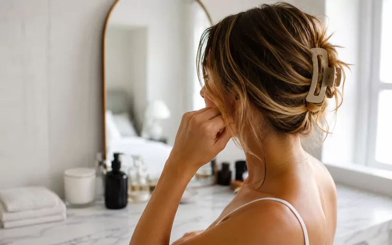 Woman styling her hair in a claw clip while standing in a bright bathroom with natural light.