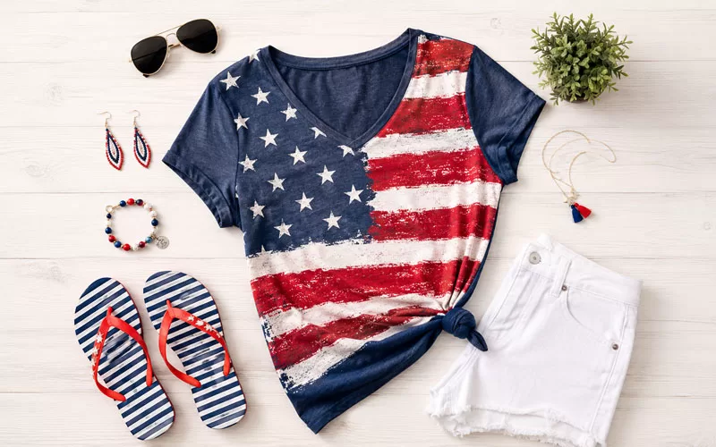 Patriotic summer outfit with an American flag shirt, white shorts, striped sandals, sunglasses, earrings, and red white and blue accessories.