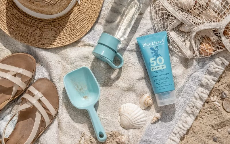Beach towel flat lay with sunscreen, water bottle, straw hat, sandals, shell bag, scoop, and seashells